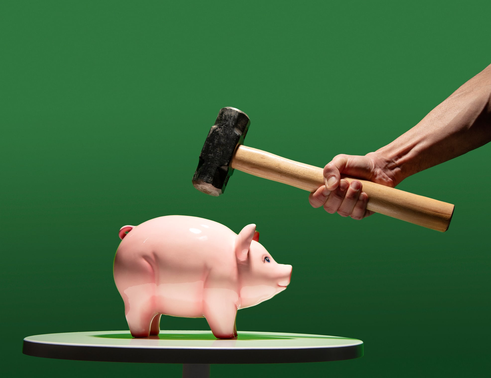 Man's hand about to hit ceramic coin piggy bank with sledge hammer, bank on white table, green back ground.