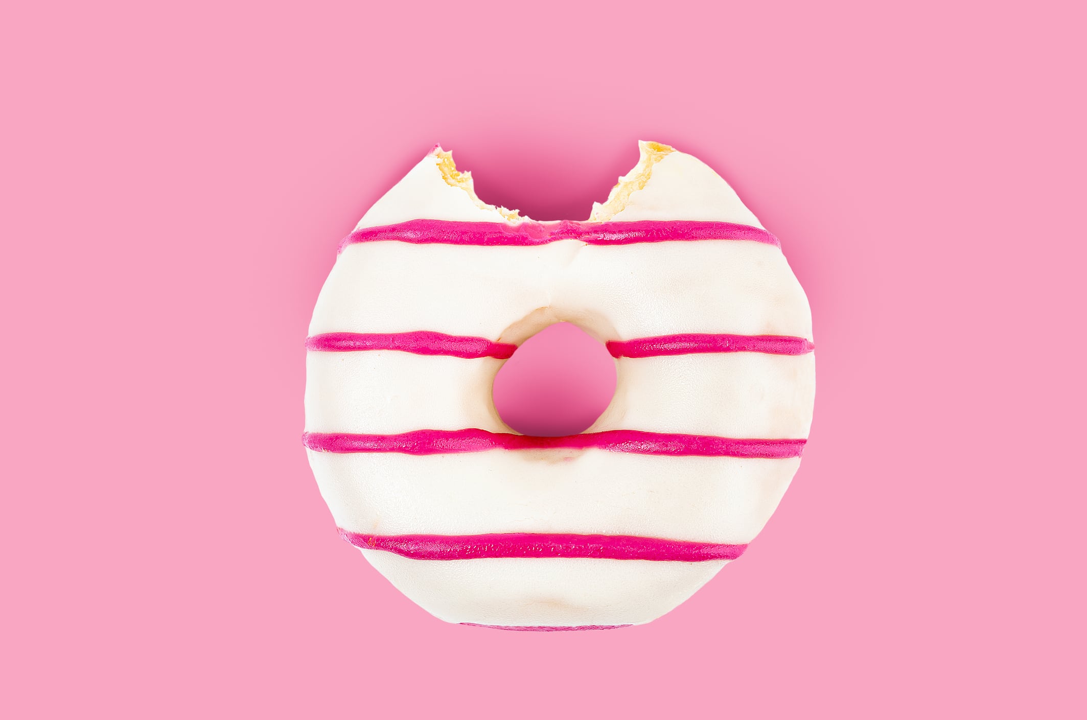 One bite missing of pink striped and white chocolate donut on pink background.