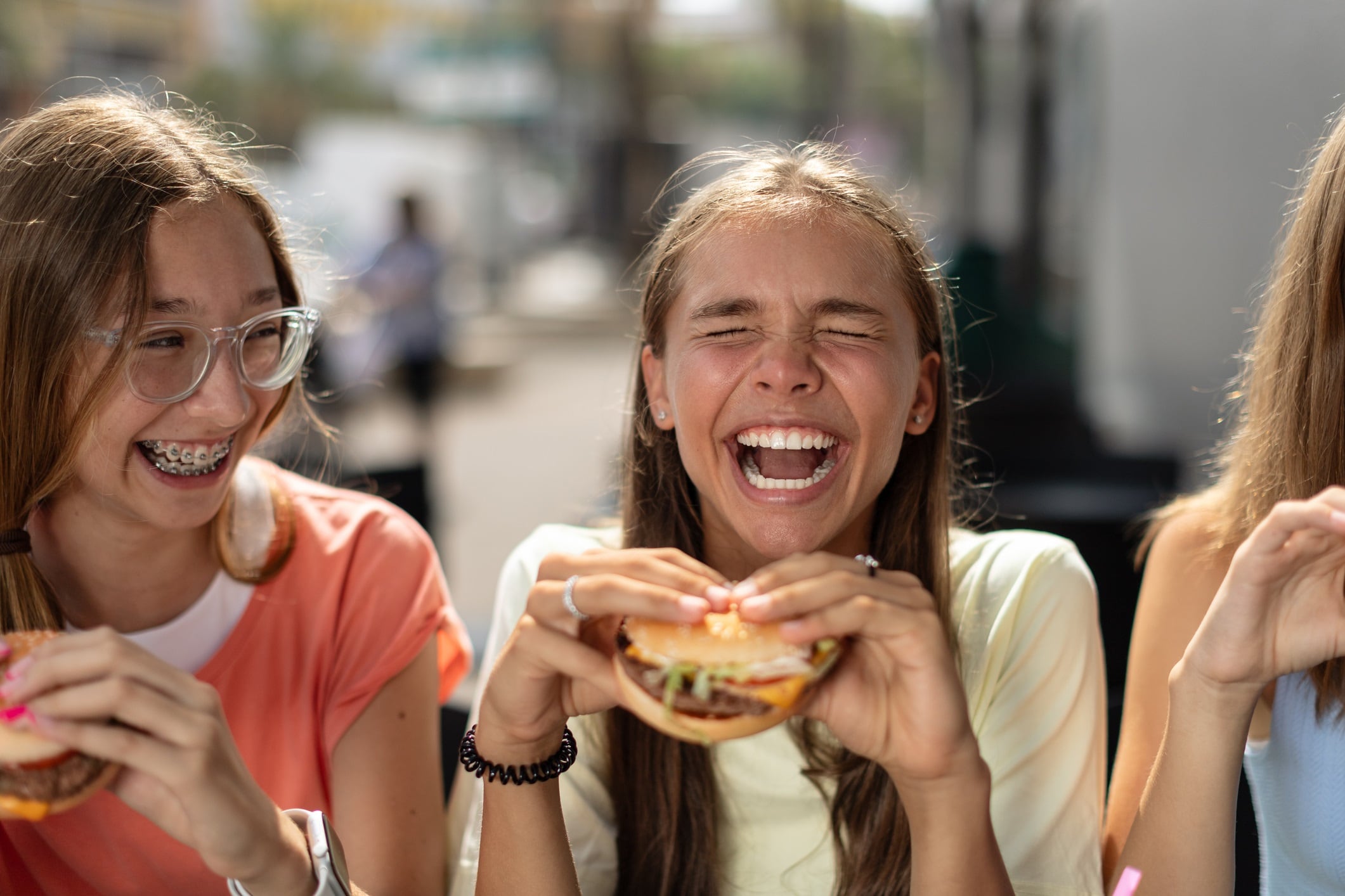 Smiling teenage girls (14-15 years old) sitting at a cafe outdoors, enjoying hamburgers.