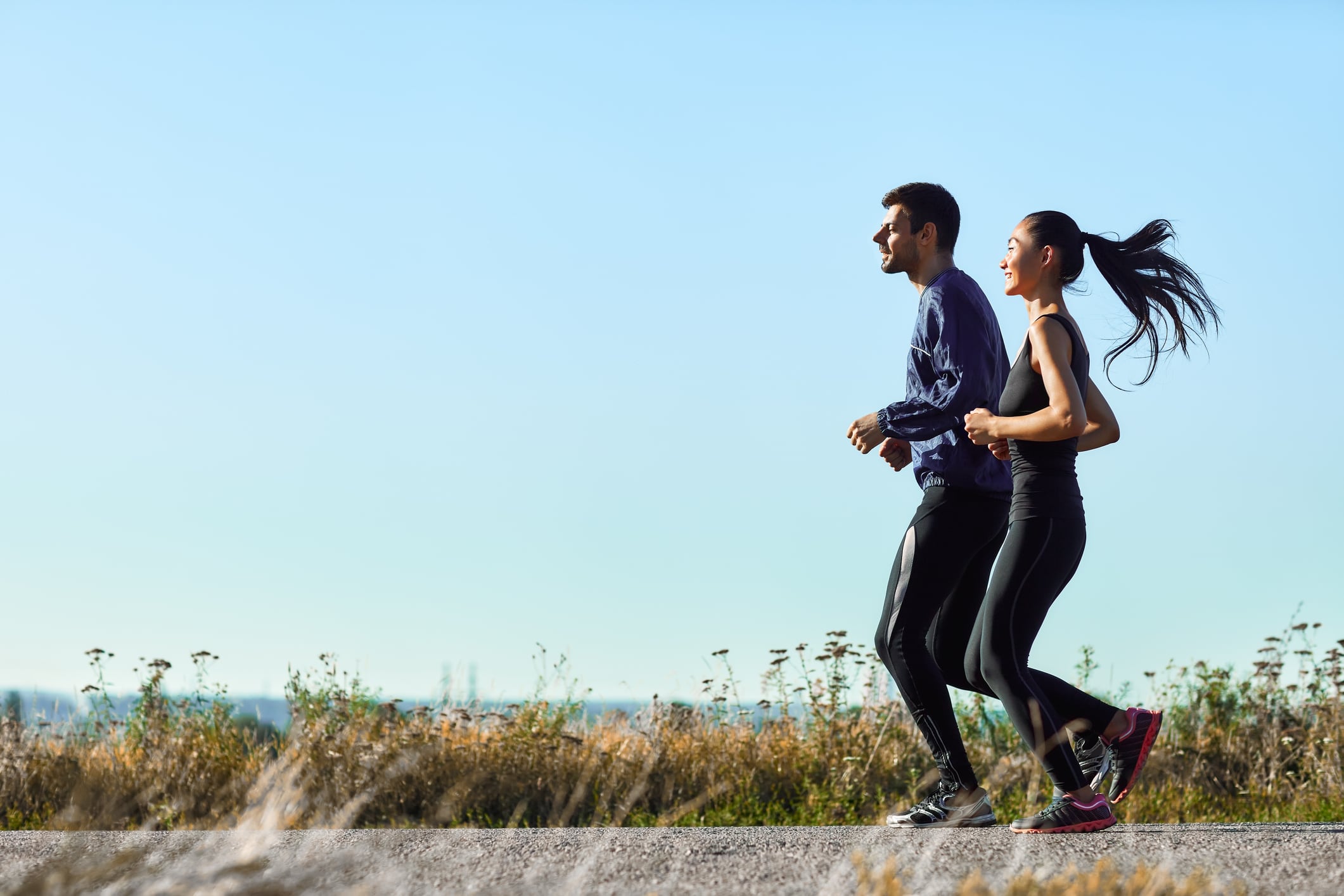 Young woman and man running along rural road. Athletic girl and guy in sportswear on morning jog outdoors. Photo with copy space on blue sky background. Full-length portrait of two sports people
