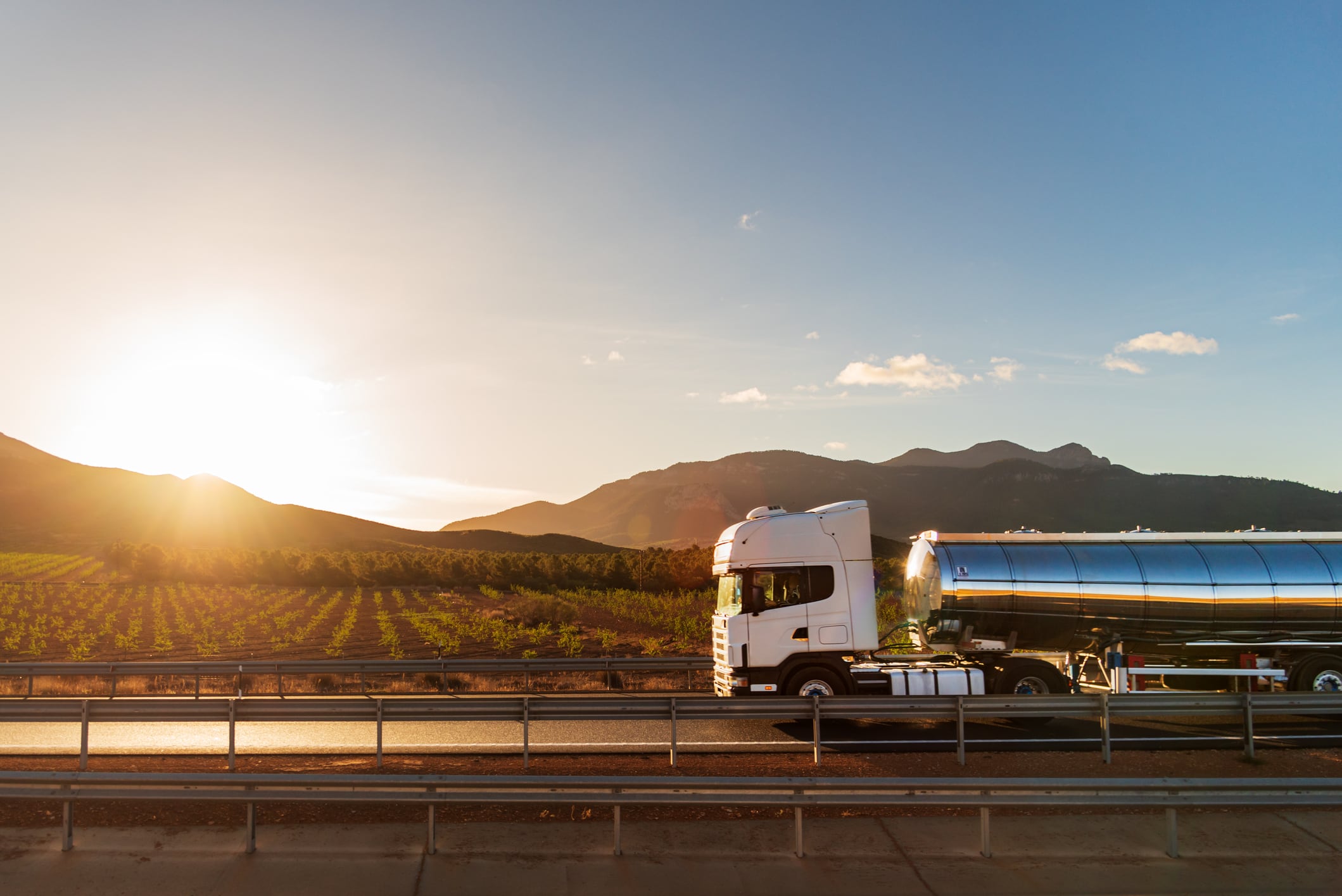Truck with a food tanker driving next to a field of vineyards with the sun rising between the mountains.