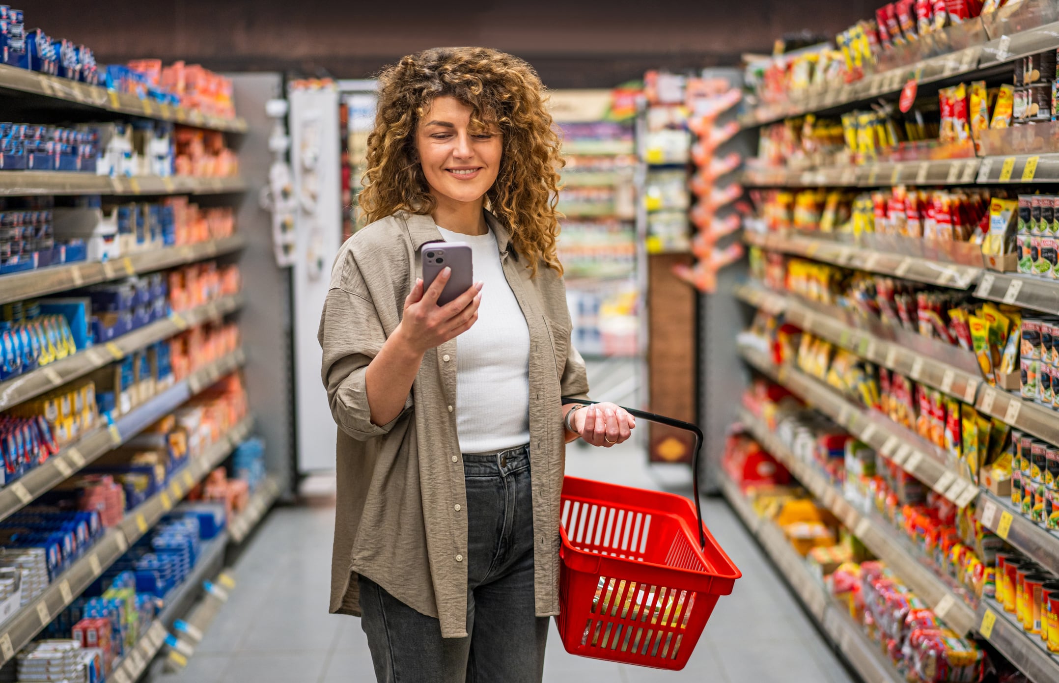 Smiling woman holding a shopping basket and using a smartphone while navigating through supermarket aisles, checking her shopping list and comparing prices