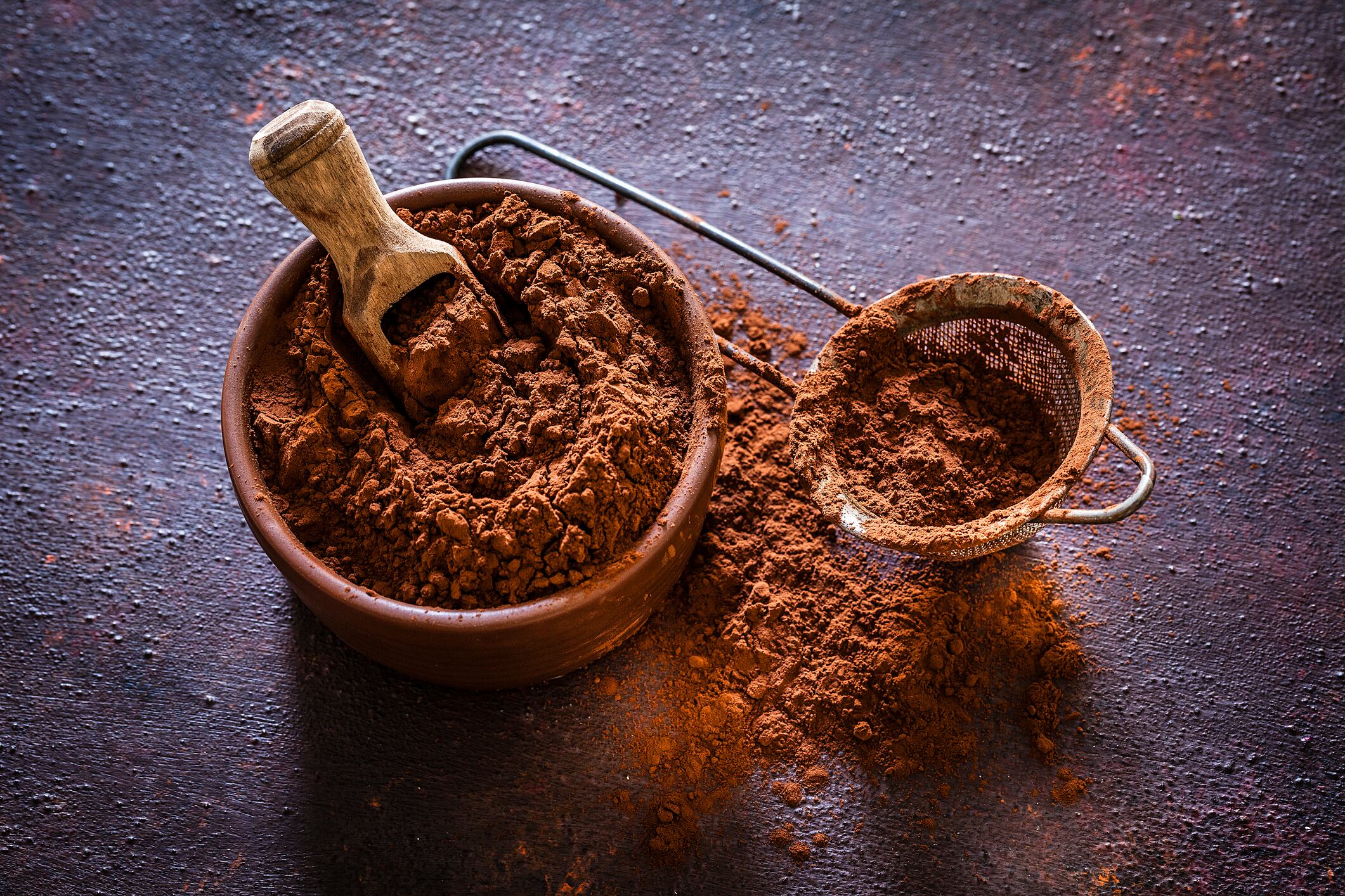 Image: Top view of a brown bowl filled with cocoa powder shot on rustic brown table. Beside the bowl is a sieve also with cocoa powder. Predominant color is brown. Low key DSRL studio photo taken with Canon EOS 5D Mk II and Canon EF 100mm f/2.8L Macro IS USM.