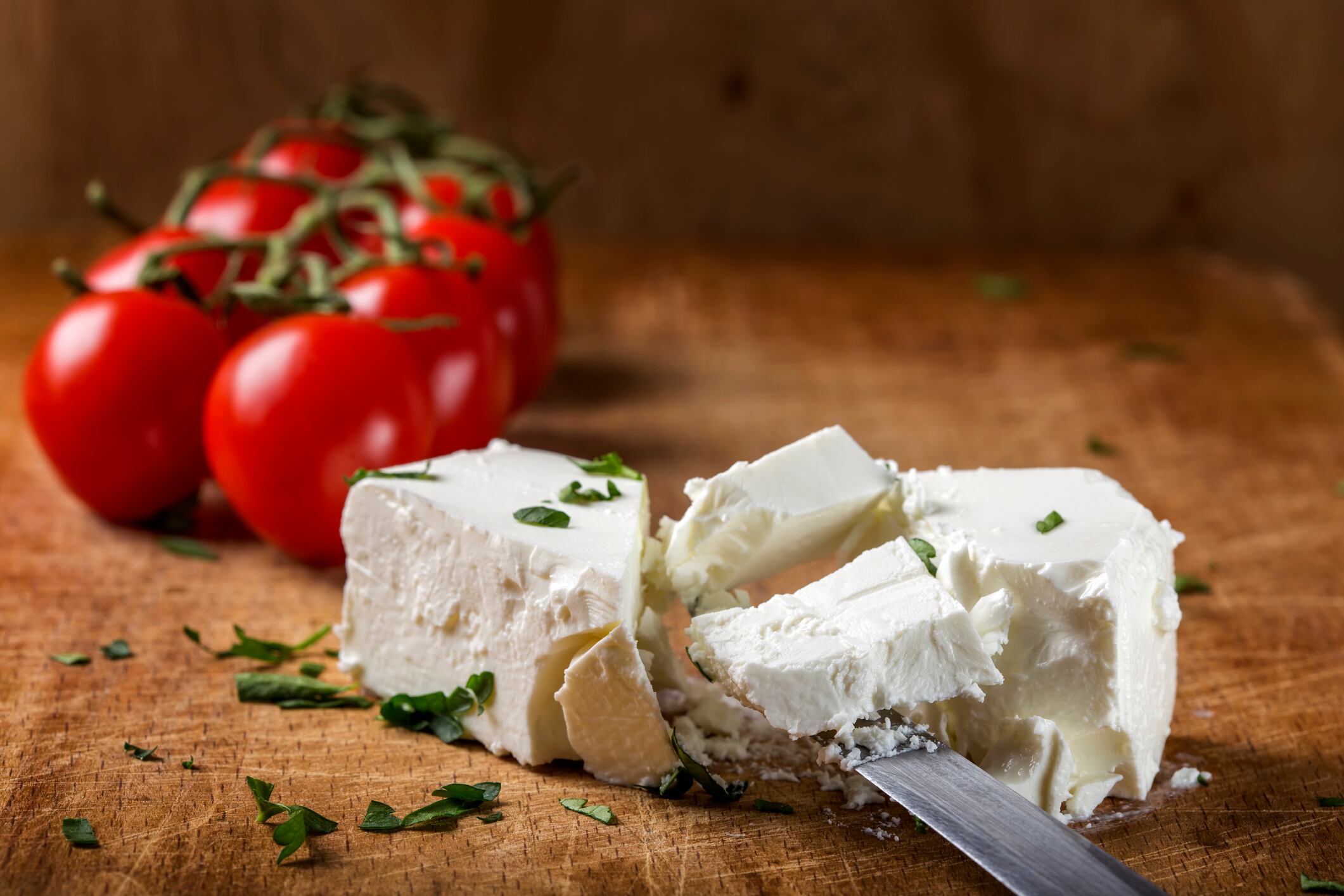 Feta cheese with knife and bunch of cherry tomatoes in background on wood with herbs