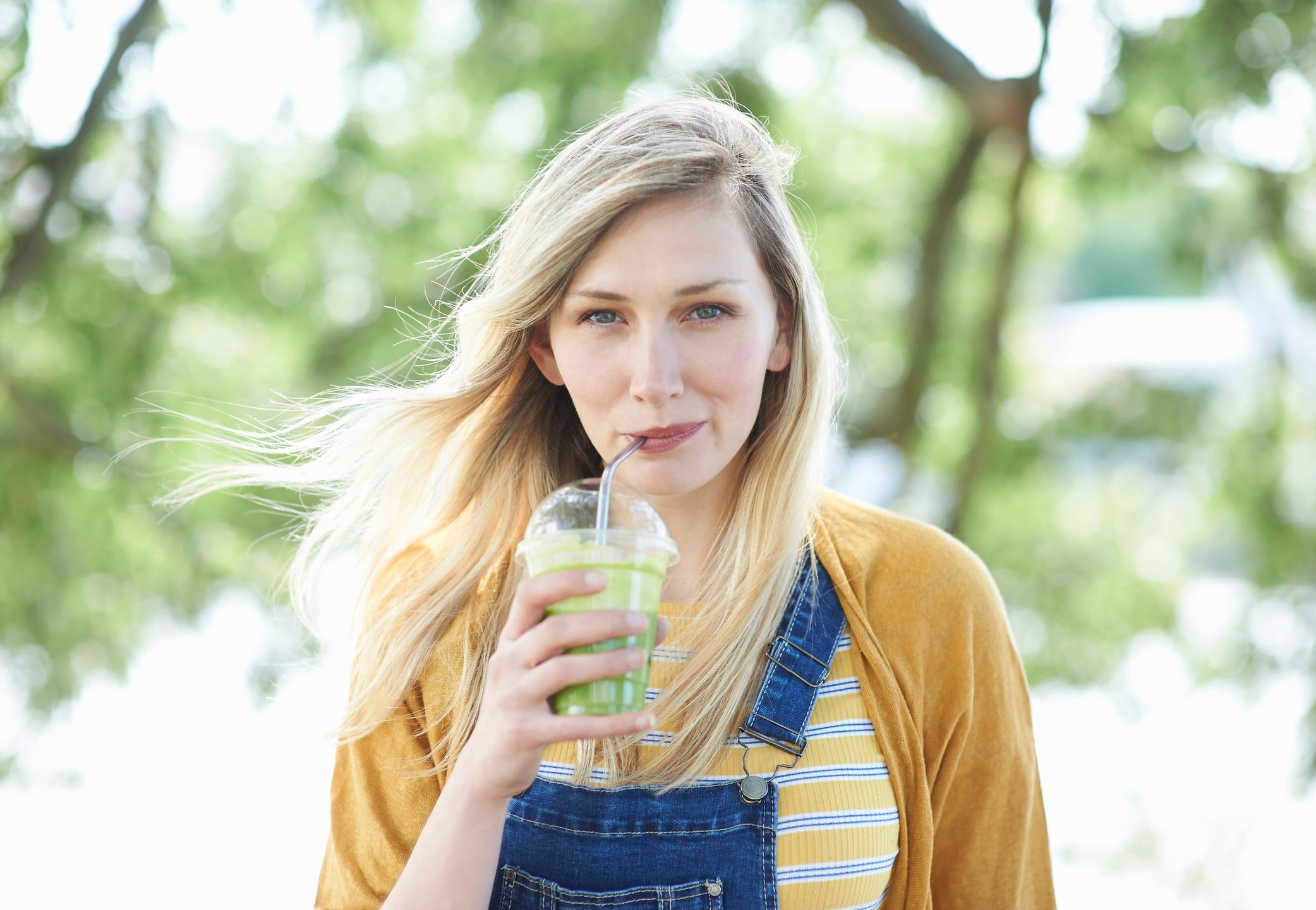 A young woman standing in the countryside drinking a freshly made fruit smoothie from a plastic free compostable cup and reusable stainless steel straw.