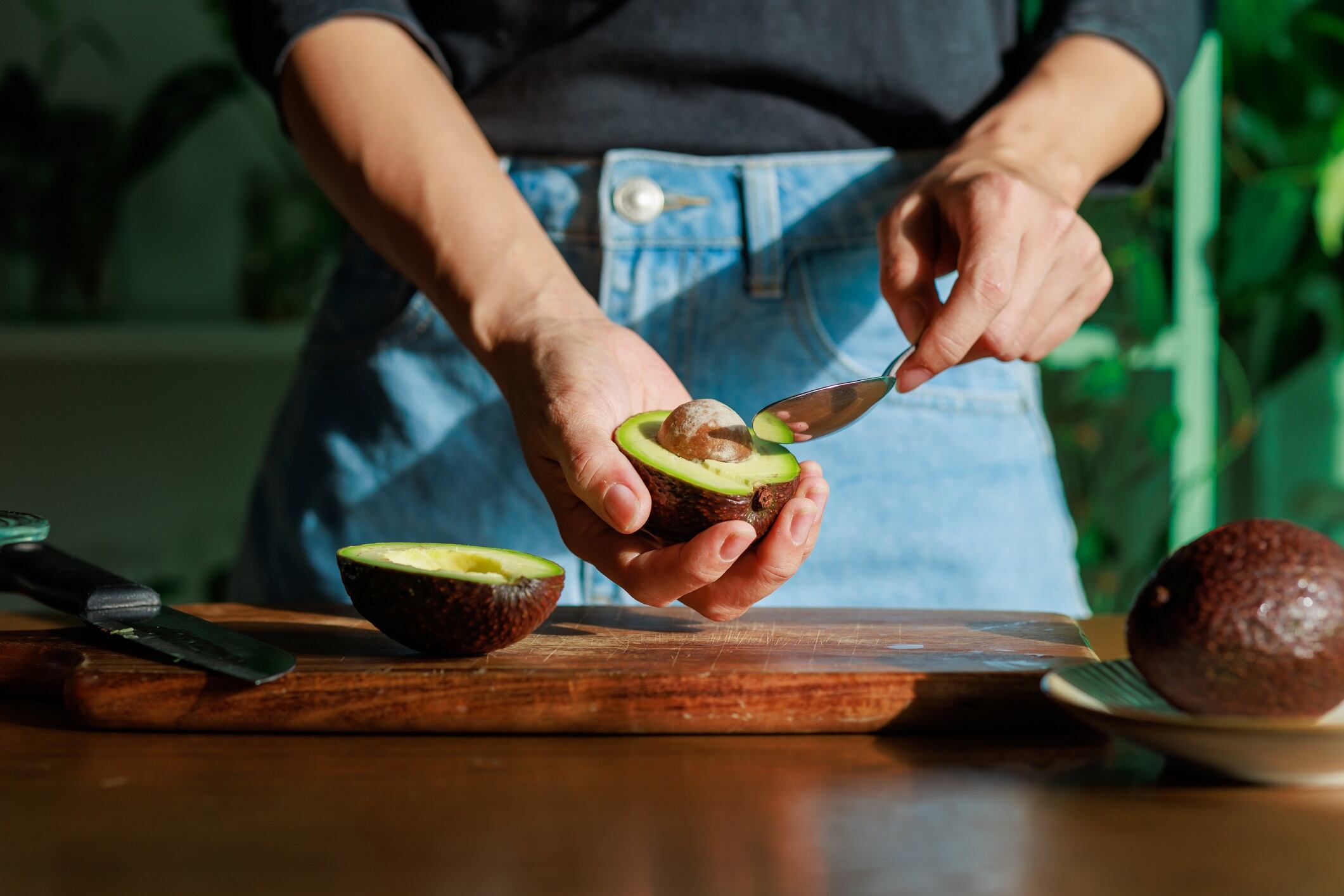 A close-up photograph capturing a woman skillfully peeling an avocado with a spoon on a cutting board, with the beautiful morning sun shining on the avocado.