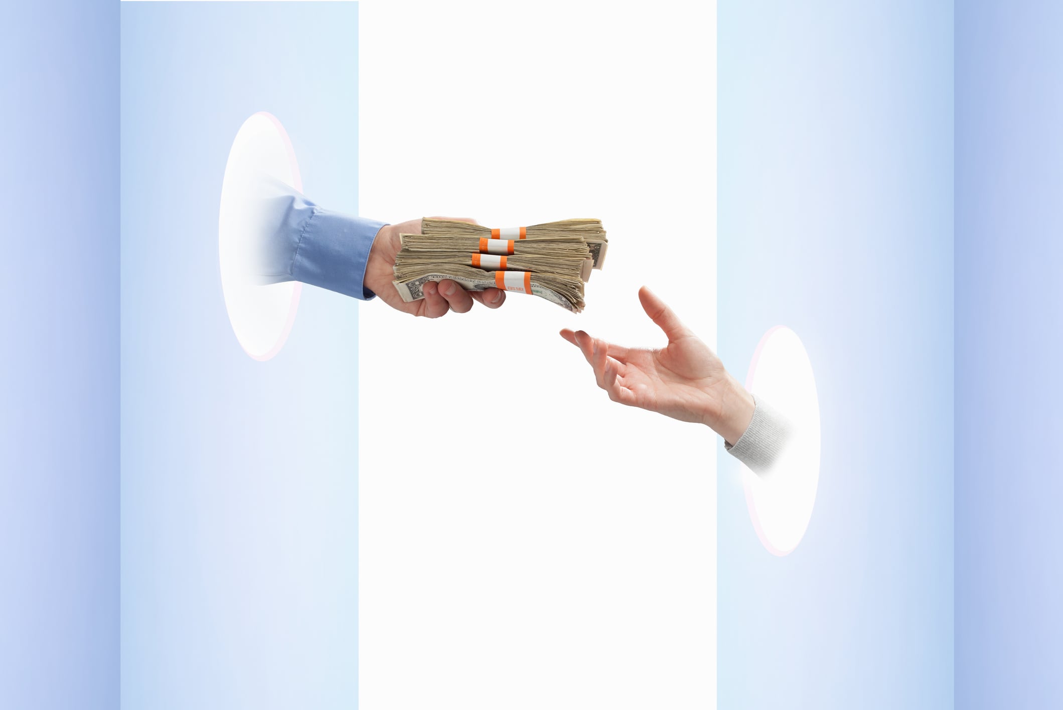 Man's hand holding bundles of US paper currency coming through hole on blue wall, woman's hand reaching in from opposite side to take money
