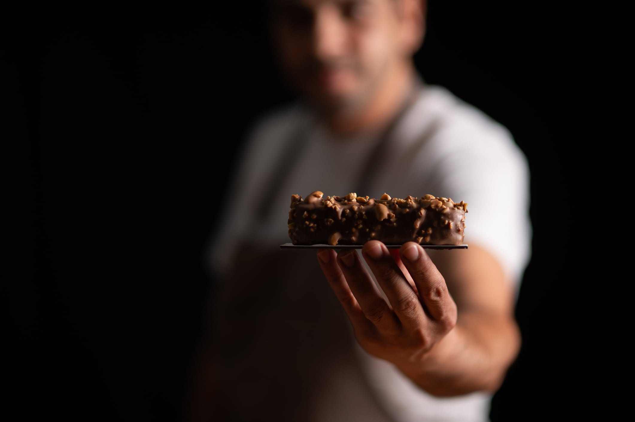 A male pastry chef holding a chocolate covered peanut cake on dark background.