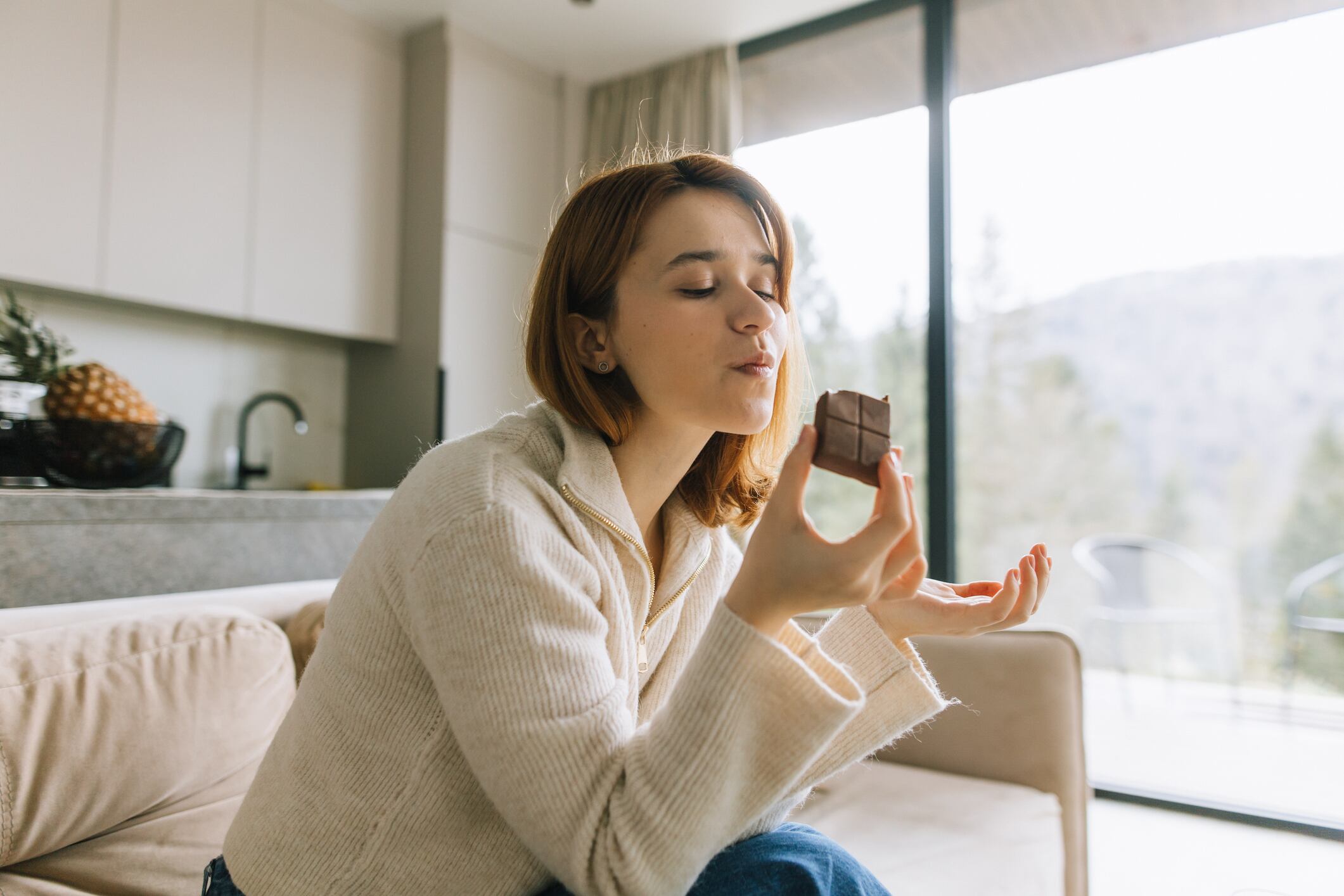 Mindful indulgence in confectionery is growing. Image: A young, beautiful woman sits comfortably on her plush sofa, fully immersed in the delightful experience of tasting a piece of rich, creamy chocolate