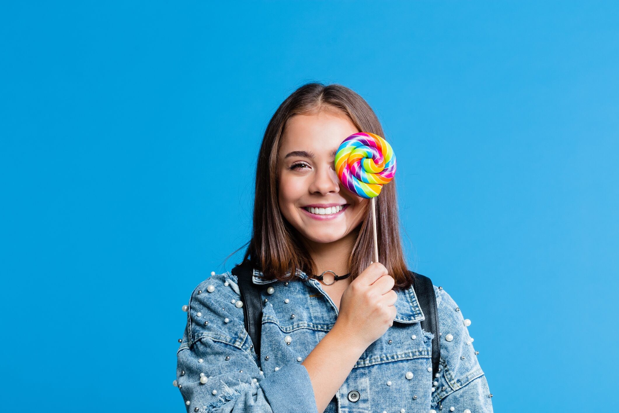 Girl wearing oversized denim jacket, standing against blue background, and  holding colourful lollipop over one eye.