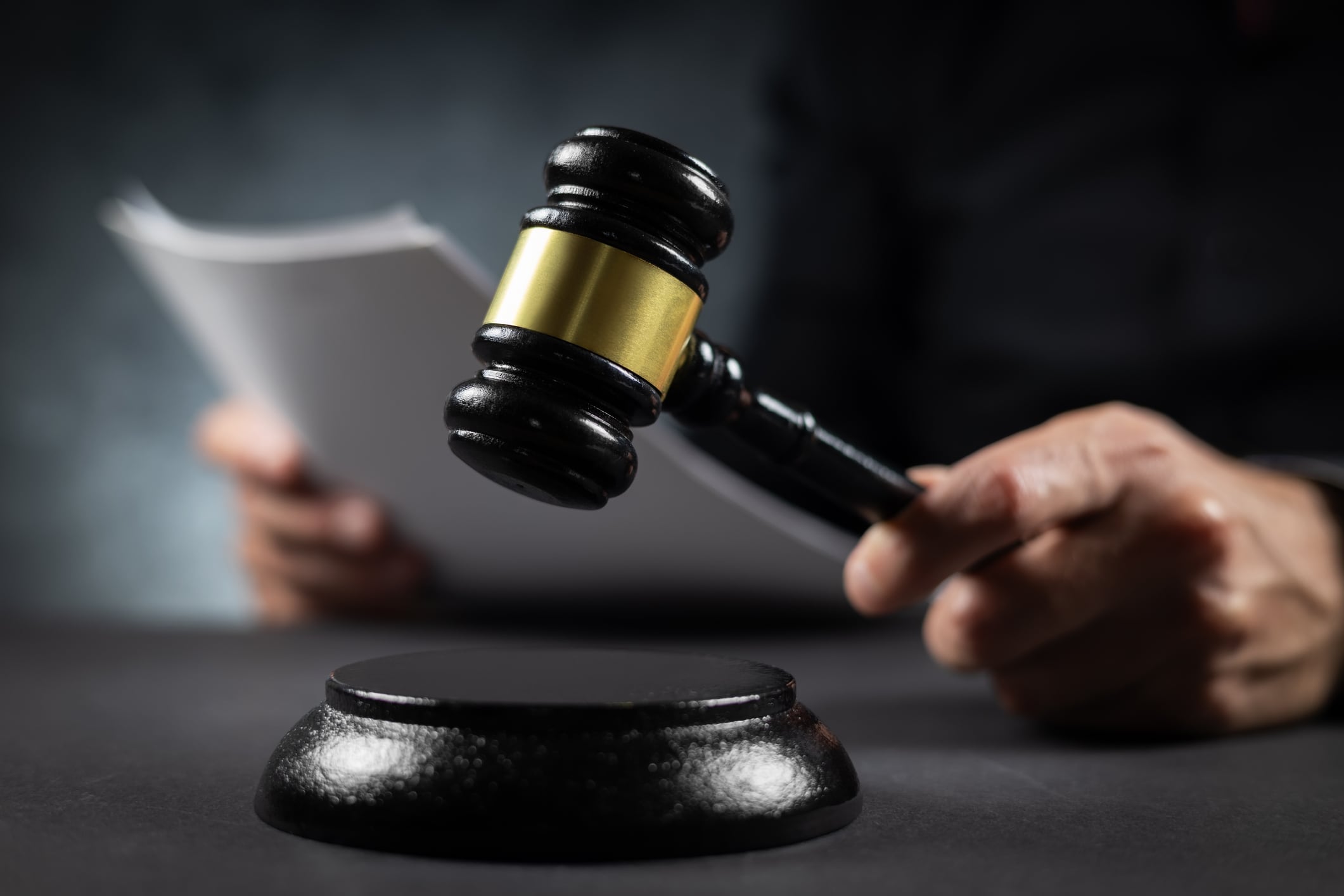 Judge with paper document pronouncing sentence in a court of law. Judge finds the accused guilty, passes judgement and rules case closed. Hand holding gavel and hitting sound block in close-up