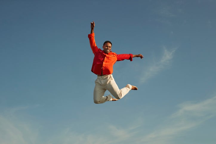 Portrait of smiling man jumping with hand raised against sky