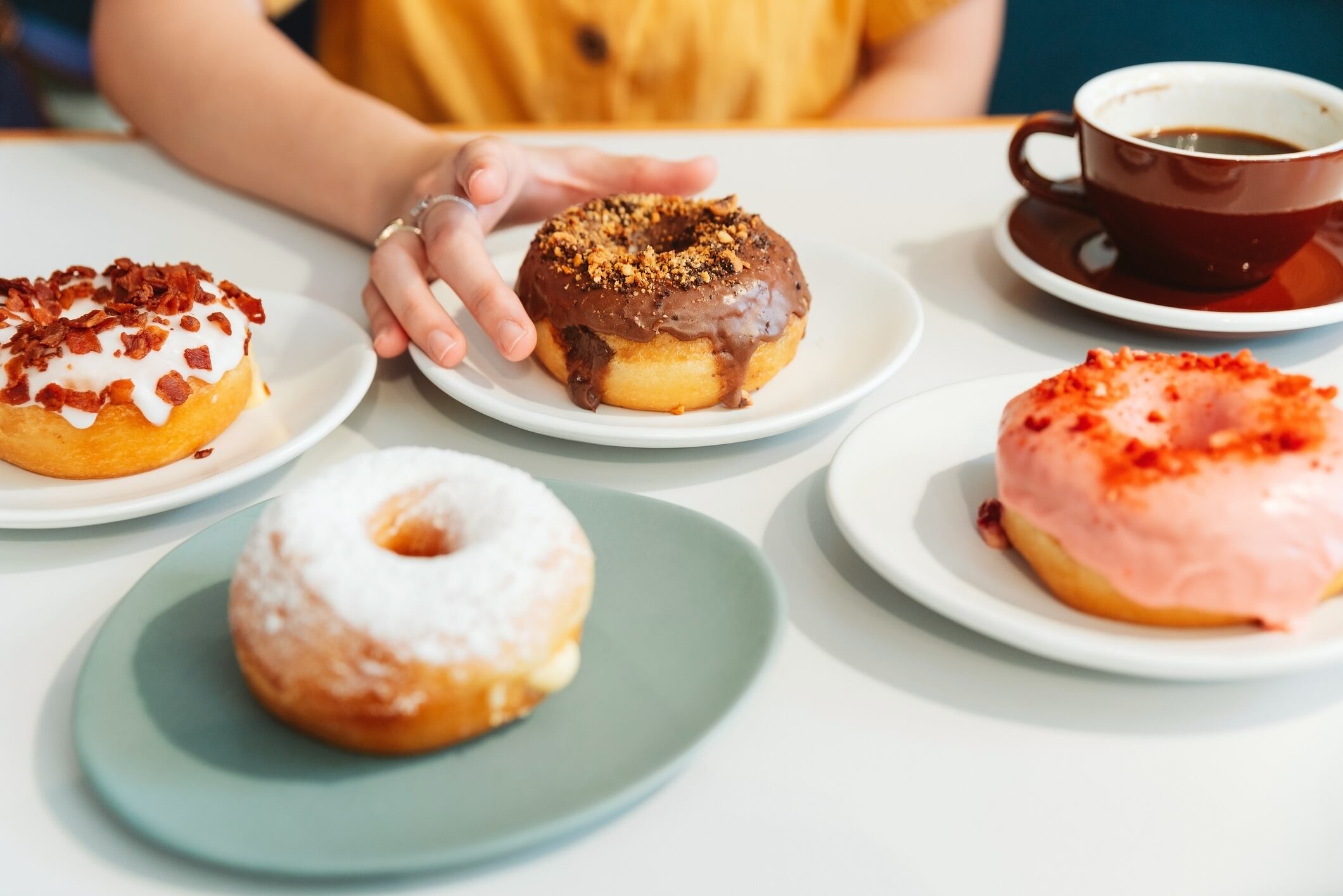 Donuts on the table include Strawberry pink, chocolate, sugar glazed and bacon, cheese donut on the table with the woman wearing yellow shirt in background in modern cafe. Enjoyment female lifestyle.
