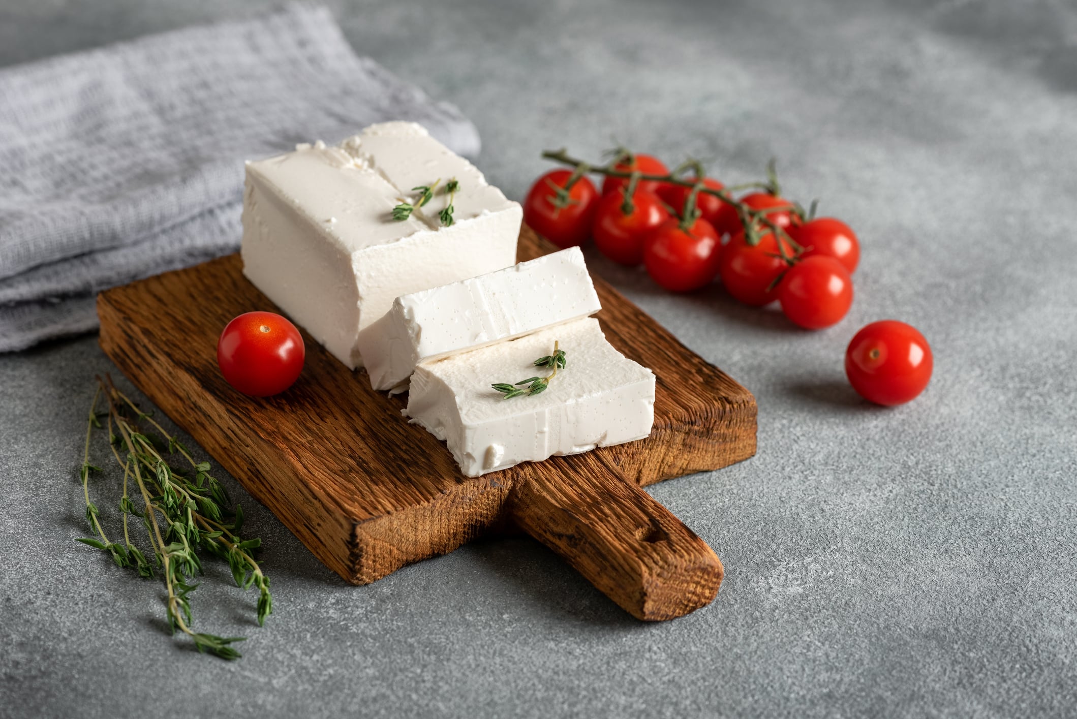 Fresh feta cheese on a wooden board with thyme and cherry tomatoes, gray concrete background. Side view, selective focus.