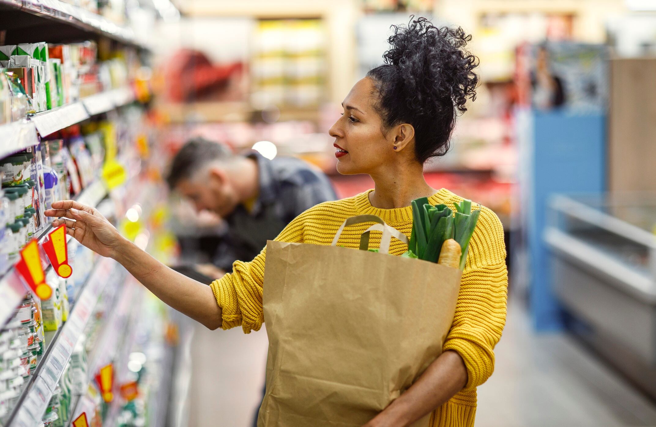 A woman, dressed in a yellow sweater, selects products in a grocery store, carrying a paper bag with fresh vegetables peeking out.