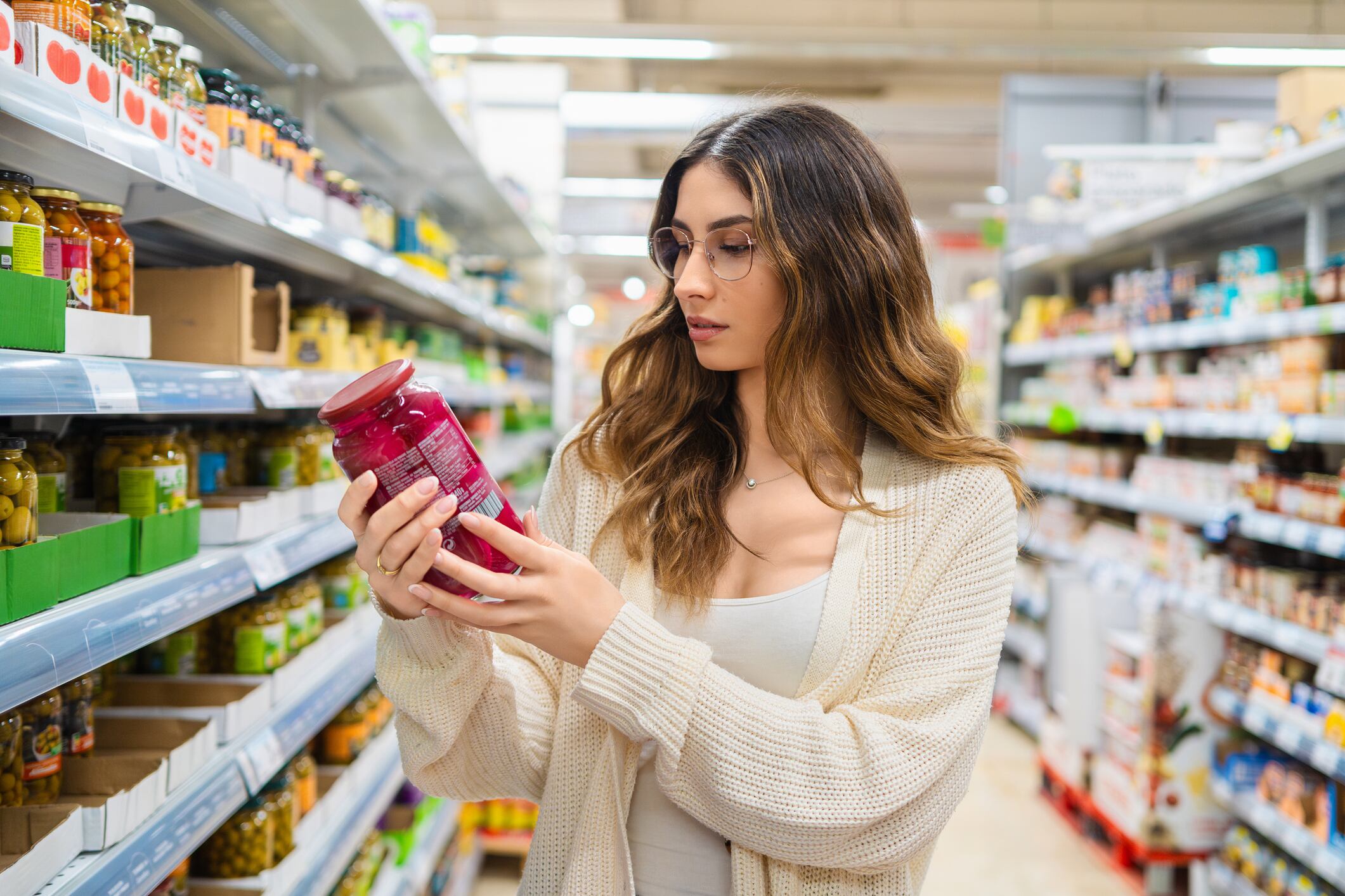 Young adult woman with glasses examining a jar in a grocery store aisle. She is focused on reading the label, surrounded by a variety of food products