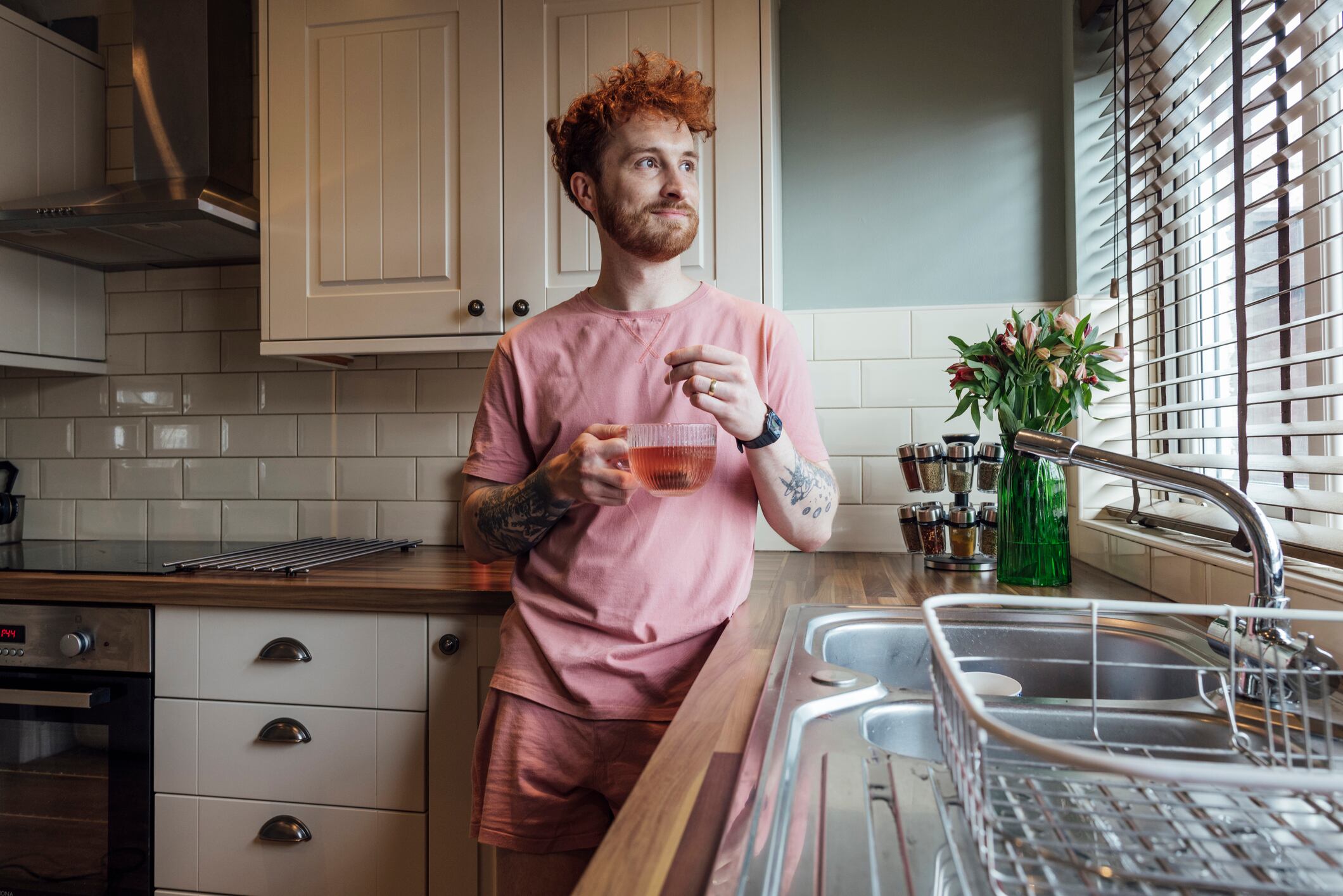 Three quarter length of a young redhead man standing in his kitchen on a morning, he is holding a cup of herbal tea and looking out the window at his home in County Durham, England. He's wearing a casual top and shorts.