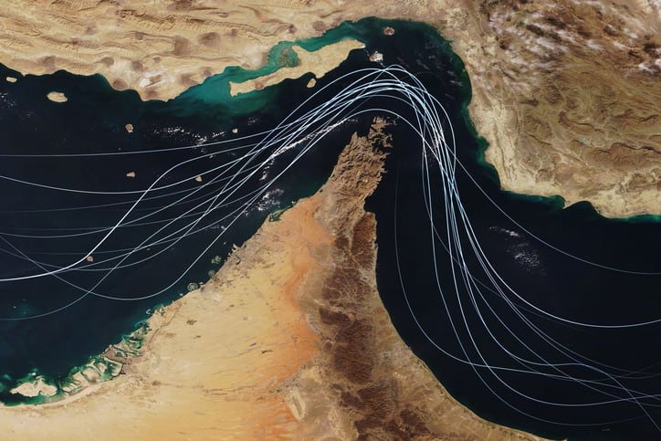 Satellite view of the Strait of Hormuz with white graphic lines representing global shipping lanes and maritime traffic between the Persian Gulf and Gulf of Oman. Strategic oil transport concept
