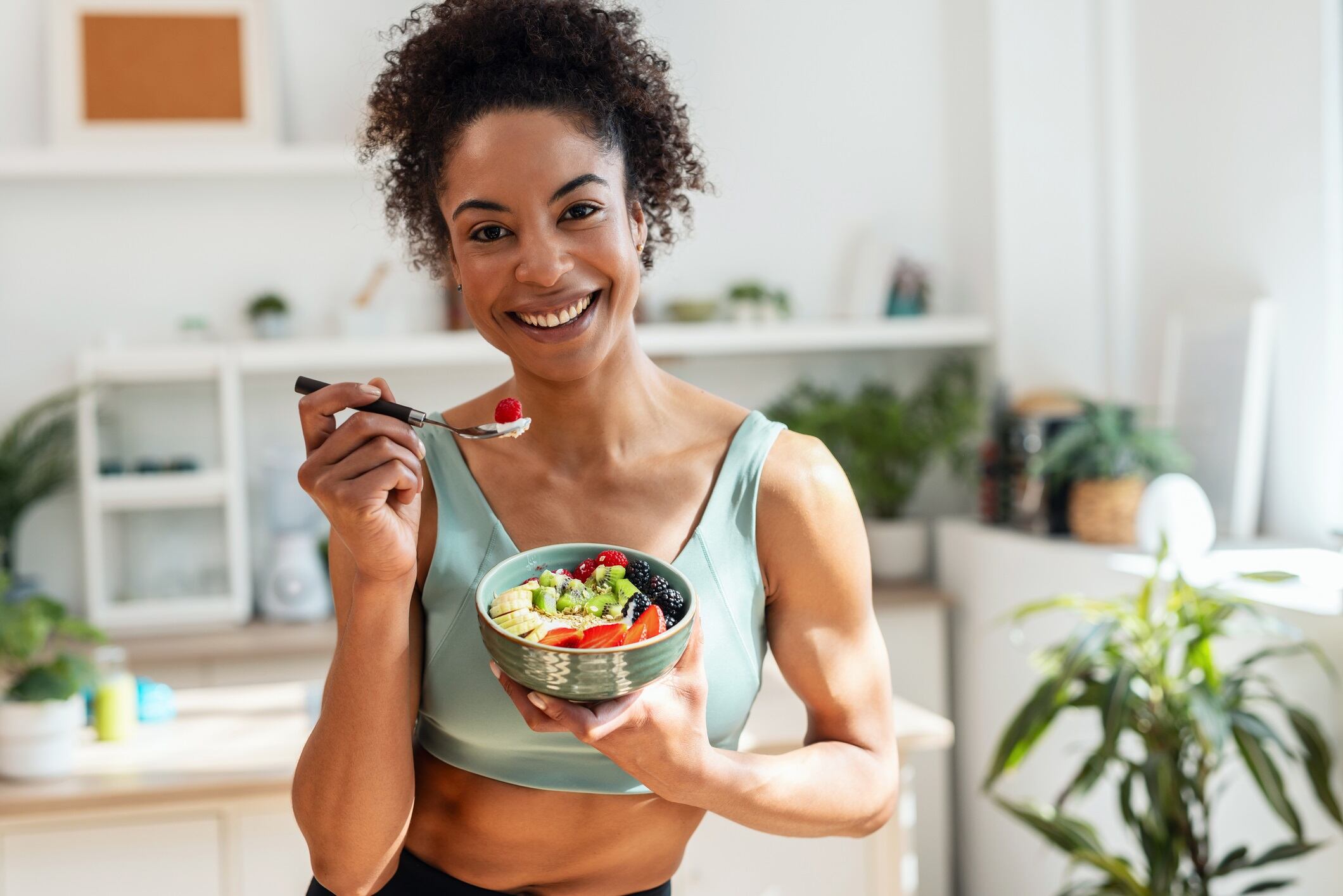 Athletic woman eating a healthy fruit bowl while looking at camera in the kitchen at home.