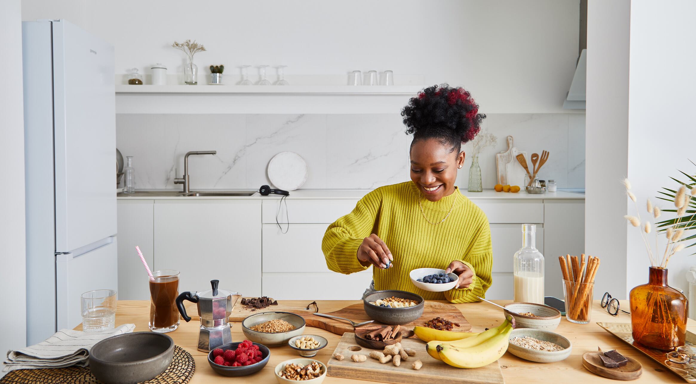 Young adult girl making oatmeal for breakfast, with fresh organic fruit, nuts and grains, at a wooden table in a modern white kitchen, representing a healthy lifestyle, an image with a copy space