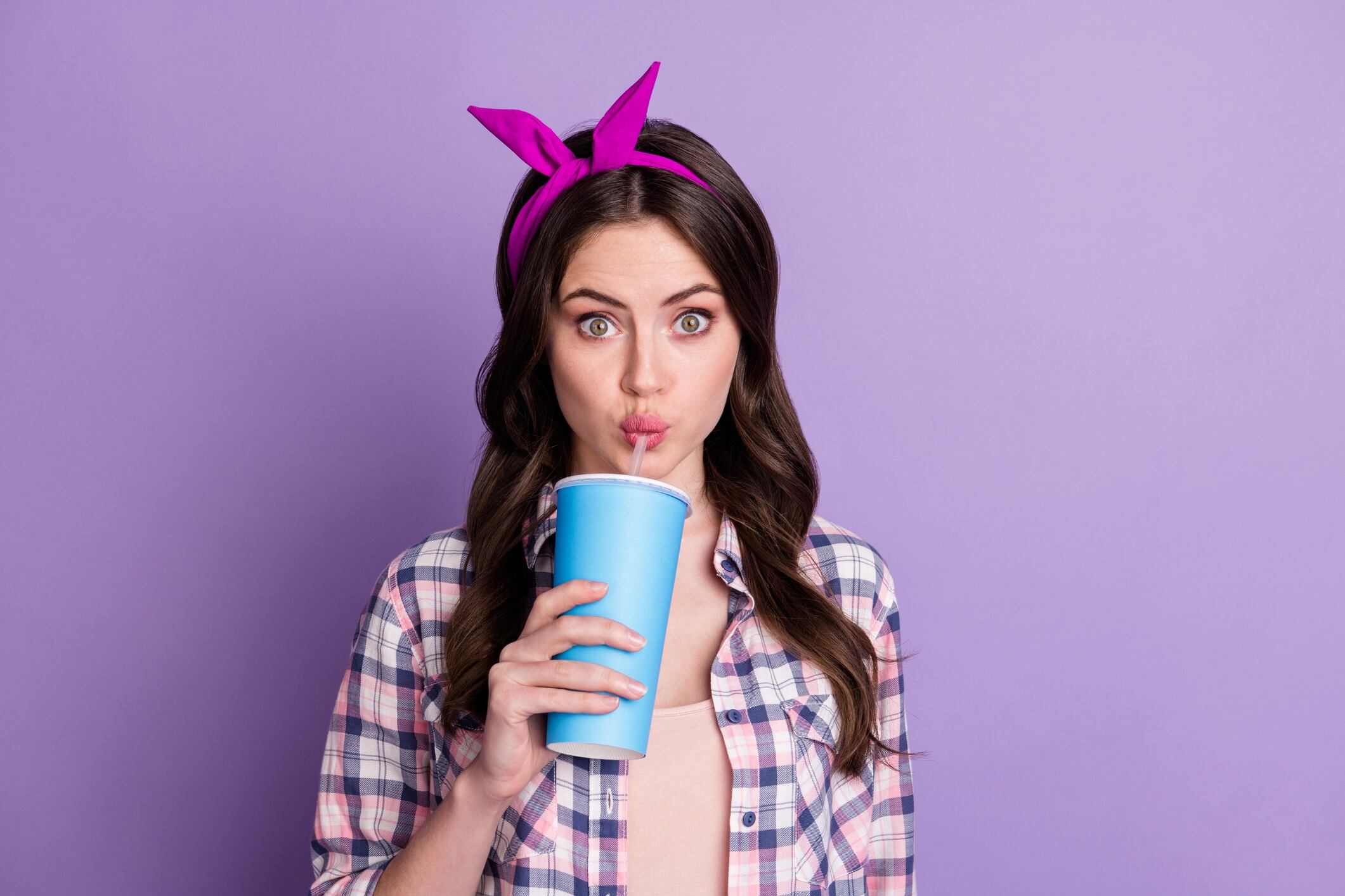 Photo portrait of girl drinking cola with plastic straw holding cup in one hand isolated on vivid violet colored background.