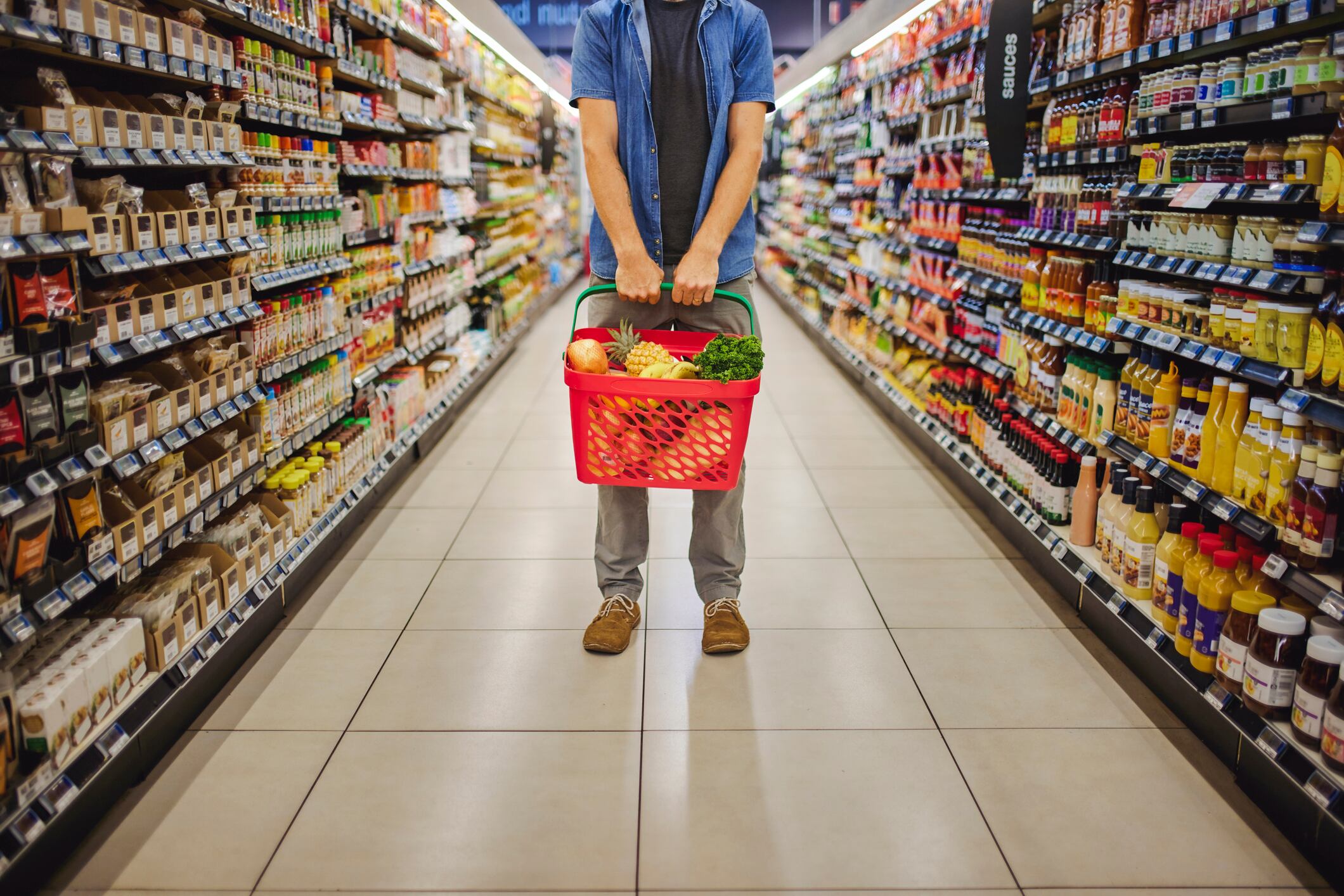 Man holding a red shopping basket filled with fresh produce while standing in a well-stocked supermarket aisle. Retail, grocery shopping, consumer behavior, aisle merchandising and product placement.